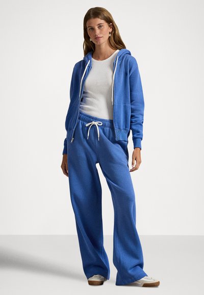 Camisola com capuz azul e calças largas combinando, feitas de tecido suave; com cintura ajustável, detalhes em branco e punhos canelados. Usada sobre um top branco.