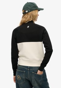 Sweat-shirt noir et crème avec un col rond, des poignets côtelés et un logo au dos. Porté avec un jean en denim bleu présentant des poches à rabat.