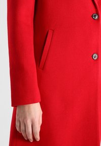 Rød uldfrakke med lige snit, udstyret med en enkelt knap, en sidelomme og en glat tekstur. Ærmeslutningerne er let tilpassede.