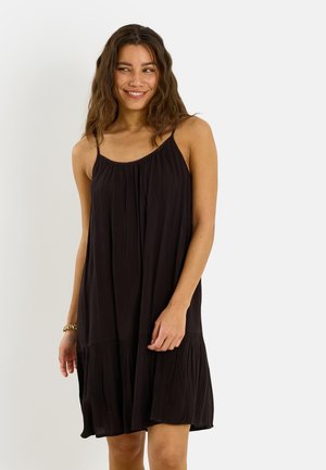 Mujer con cabello ondulado hasta los hombros, con un vestido negro holgado sin mangas, sonriendo y mirando hacia un lado sobre un fondo liso.