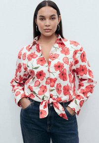 Camicia a maniche corte con bottoni, floreale, su sfondo bianco, caratterizzata da grandi motivi di papaveri rossi, annodata in vita, abbinata a jeans blu scuro.