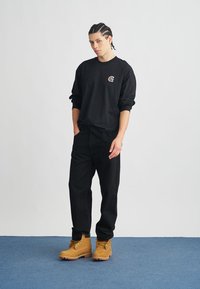 Black long-sleeve shirt with logo, black pants, and tan boots. The outfit features a relaxed fit and casual, durable materials.