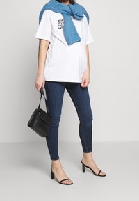 Camiseta blanca de gran tamaño con texto negro, chal azul de punto alrededor de los hombros, jeans ajustados azul oscuro, bolso negro y sandalias de tacón negras.