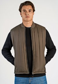 Brun vadderad väst med vertikal söm, dragkedja fram och ribbstickade svarta ärmar. Modellen står med händerna i fickorna.