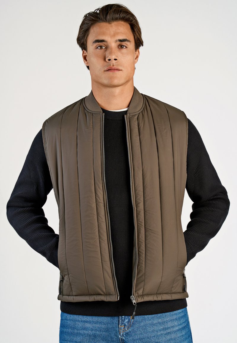 Brun vadderad väst med vertikal söm, dragkedja fram och ribbstickade svarta ärmar. Modellen står med händerna i fickorna.