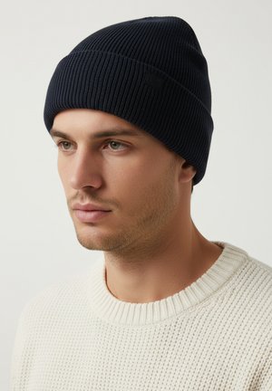 Junger Mann mit leichtem Stoppelbart, der eine schwarze Strickmütze und einen weißen, strukturierten Pullover trägt, schaut leicht zur Seite vor neutralem Hintergrund.