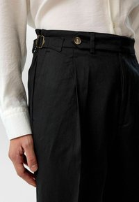 Zwarte op maat gemaakte broek met een gestructureerde afwerking, met voorplooien, een riem met een gouden gesp en een contrasterende knop bij de taille.