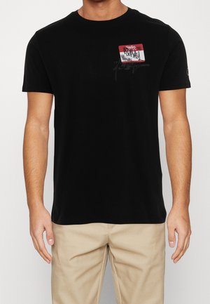 Mand iført en sort T-shirt med korte ærmer, dekoreret med et rødt og hvidt "Hej, mit navn er" klistermærke på brystet, parret med beige bukser.