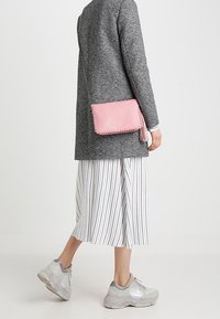 Borsa a tracolla in pelle rosa con dettagli in cordoncino intrecciato e accentuazione con frange, indossata sopra un cappotto grigio e una gonna a righe bianche.