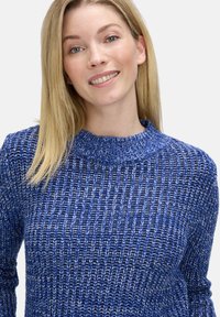 Pull en tricot bleu avec un col montant, un motif texturé et des poignets côtelés. Le tissu présente des nuances variées de bleu et de blanc.