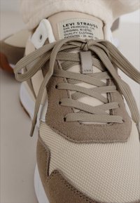 Sneaker beige e tortora con una parte superiore texture, che presenta sezioni in camoscio e tessuto, lacci tondi e un'etichetta sul lato. Suola in gomma.