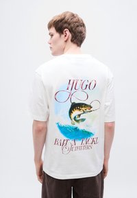 Λευκό βαμβακερό t-shirt με πολύχρωμο γραφικό ενός ψαριού σε κύμα και το κείμενο "HUGO" και "BAIT 'N TACKL OUTFITTERS" από κάτω.