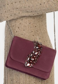 Bordowa zamszowa torebka na ramię z zakrzywioną ozdobą z kryształów, srebrnym łańcuszkiem i gładkim zamknięciem na klapę. Teksturowane tło materiału.