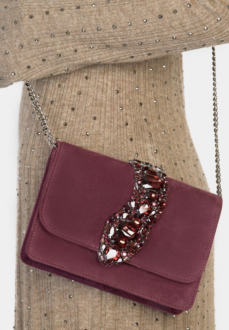 Bordowa zamszowa torebka na ramię z zakrzywioną ozdobą z kryształów, srebrnym łańcuszkiem i gładkim zamknięciem na klapę. Teksturowane tło materiału.