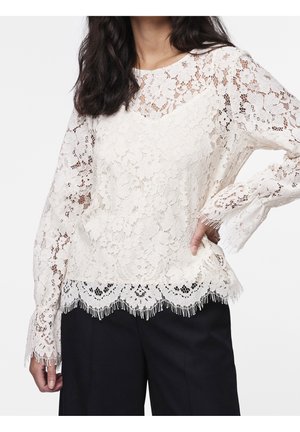 Kobieta ubrana w białą, kwiatową koronkową bluzkę z długim rękawem i falistym wykończeniem, w połączeniu z czarnymi spodniami, pozująca z jedną ręką na biodrze.