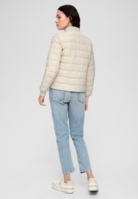 Veste en duvet matelassée beige avec un col montant, dotée de coutures horizontales, associée à un jean bleu clair et des baskets blanches.