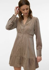 Femme aux cheveux bruns mi-longs portant une robe beige à manches longues boutonnée, à taille cintrée et ourlet à volants.