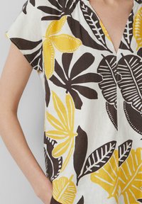 Blouse à manches courtes en tissu léger, avec un fond blanc orné de motifs tropicaux noirs et jaunes audacieux et un décolleté en V.