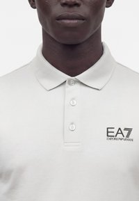 Gros plan d'une personne portant un polo blanc à trois boutons avec un logo EA7 Emporio Armani sur la poitrine.