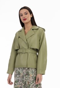 Giacca in ecopelle verde con vita cintrata, colletto ampio e grandi bottoni. Indossata sopra una gonna a fantasia con design floreali e astratti.