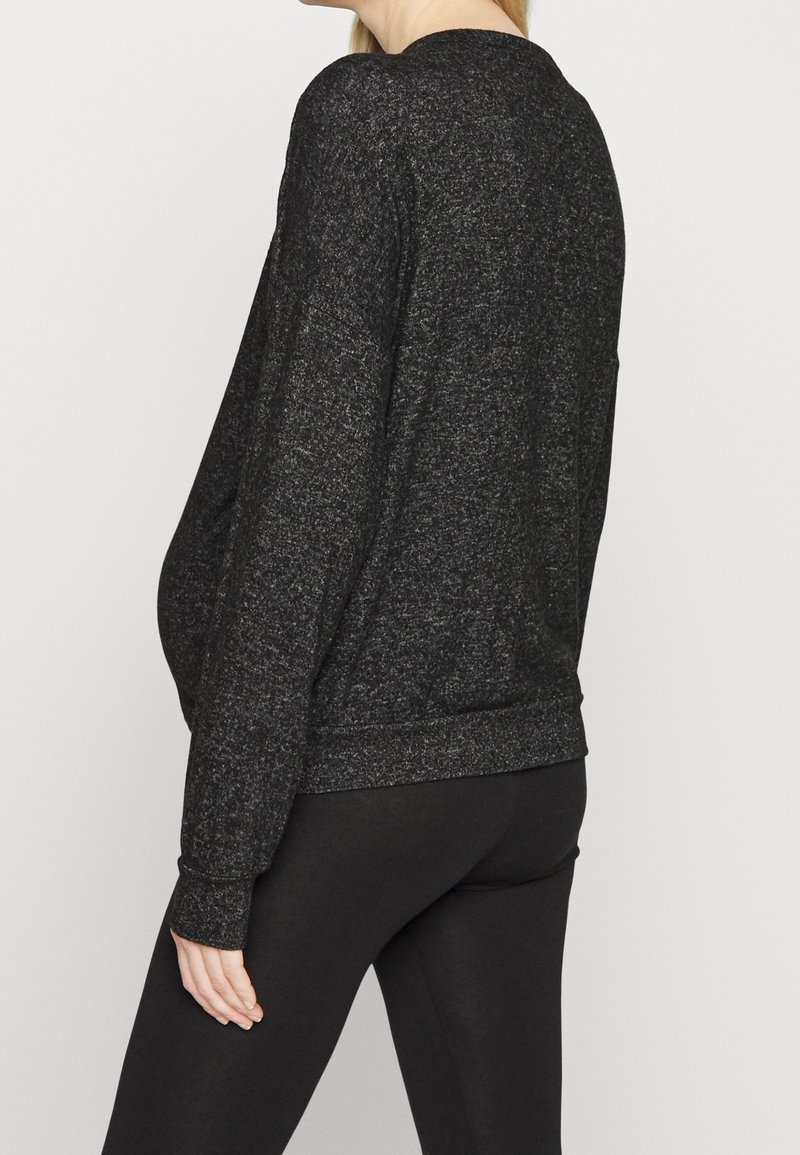 Femme portant un pull à manches longues gris foncé et des leggings noirs, debout de profil sur un fond clair uni.
