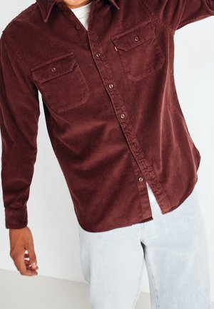 Mann trägt ein zugeknöpftes weinrotes Cordhemd mit Brusttaschen und hellblauen Jeans vor neutralem Hintergrund.