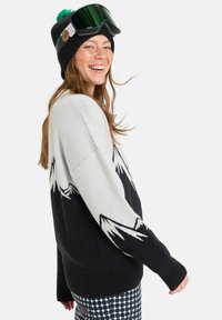Knit sweater featuring a gray body with black mountain pattern, ribbed cuffs, and a relaxed fit. Paired with a black beanie and ski goggles.
