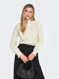 Lichtgele blouse met bloemrijke borduursels en ruches, gecombineerd met zwarte hoge taillebroek en een zwarte handtas.