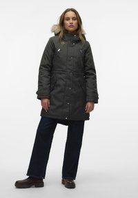 Parka verde scuro con cappuccio staccabile foderato in pelliccia, tasche con zip e taglio dritto, abbinata a jeans larghi scuri e stivali marroni.