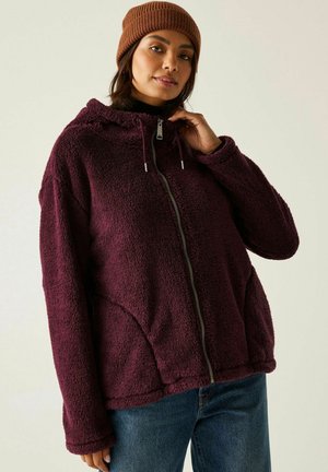 Mujer con una sudadera con capucha marrón granate de textura suave con cremallera, un gorro de punto marrón y pantalones vaqueros azules, ajustando la cremallera de la sudadera cerca del cuello.