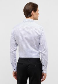 Camicia a maniche lunghe in bianco con motivo geometrico blu navy. Presenta un colletto classico e polsini con bottoni. I pantaloni neri completano il look.