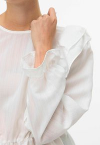 Blusa bianca con strisce verticali rosa pallido, maniche arricciate, texture morbida e polsini arricciati. Il materiale sembra leggero e arioso.