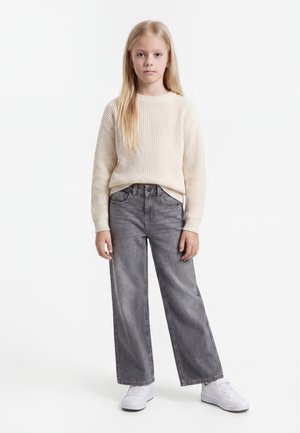 Jeune fille aux longs cheveux blonds portant un pull en maille crème, un jean gris à jambes larges et des baskets blanches, debout sur un fond blanc.