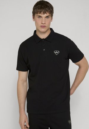 Ein junger Mann trägt ein schwarzes Poloshirt mit einem kleinen diamantförmigen Logo auf der Brust und posiert vor einem schlichten hellen Hintergrund.