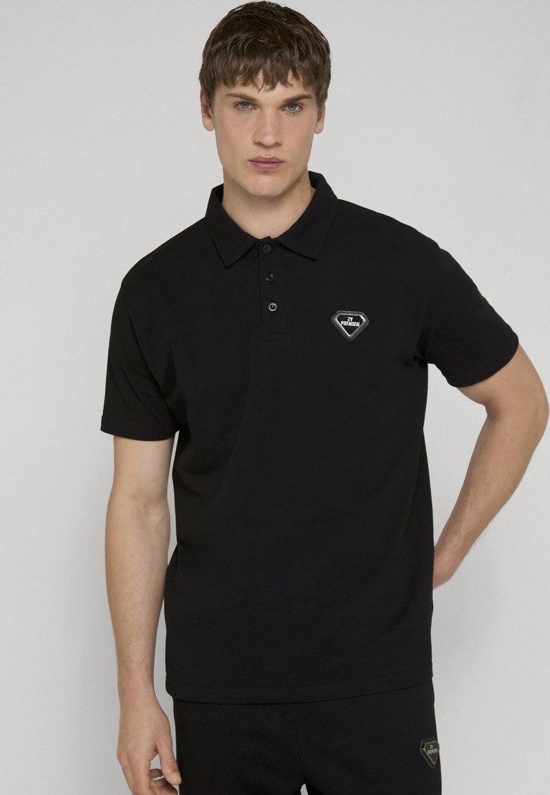 Jeune homme portant un polo noir avec un petit logo en forme de losange sur la poitrine, posant devant un fond clair uni.