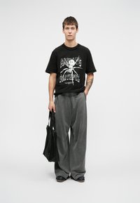 Fiatal férfi áll, kezét a zsebébe dugva, fekete grafikus pólót, laza szürke nadrágot, fekete cipőt visel, és egy fekete táska van a kezében.