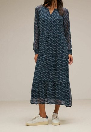 Žena v tmavě modrých midi šatech s dlouhým rukávem a vzorem, doplněných bílými nízkými teniskami, stojící na jednobarevné podlaze.