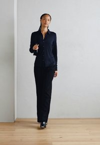 Marineblauwe overhemd met lange mouwen, gecombineerd met zwarte broek en zwarte schoenen. Model staat op een lichte houten vloer tegen een lichte muur.