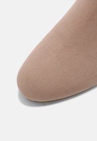 Gros plan sur la pointe d'une chaussure en daim beige à texture lisse sur un fond blanc uni.