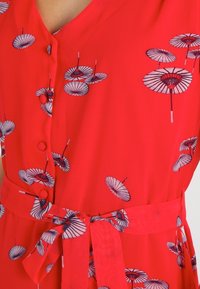 Červené šaty vyrobené z lehkého materiálu s vzorem malých deštníků. Obsahují zapínání na knoflíky vpředu a zavazovací pásek v oblasti pasu pro zvýraznění siluety.