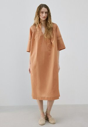 Mujer con cabello largo y ondulado que lleva un vestido suelto color terracota hasta la rodilla, con mangas hasta el codo y zapatos beige tejidos con ataduras al tobillo.