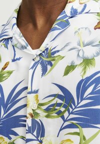 Kurzarmhemd aus weißem Stoff mit einem lebhaften Blumenmuster in Blau, Grün und Gelb. Verfügt über einen angedeuteten Kragen und eine Knopfleiste vorne.