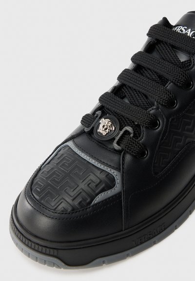 Black leather sneaker features an embossed Greek key pattern, black laces, a silver logo button, and grey accents on the side.