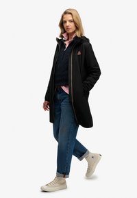 Abrigo negro con capucha, presenta detalles en naranja y cierre de cremallera, llevado sobre un suéter de punto azul marino, una camisa rosa claro y unos vaqueros. 