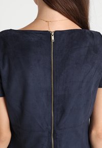 Námořnická modrá šaty z umělé suède s zapínáním na zip na zadní straně a přiléhavým střihem. Textura je hladká s mírným leskem.