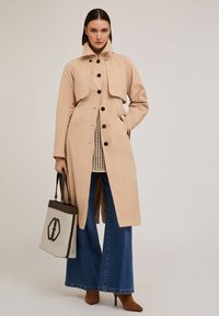 Cappotto beige con colletto alto, bottoni neri e tasche laterali. Indossato sopra un top in maglia e abbinato a jeans a gamba larga e una borsa.