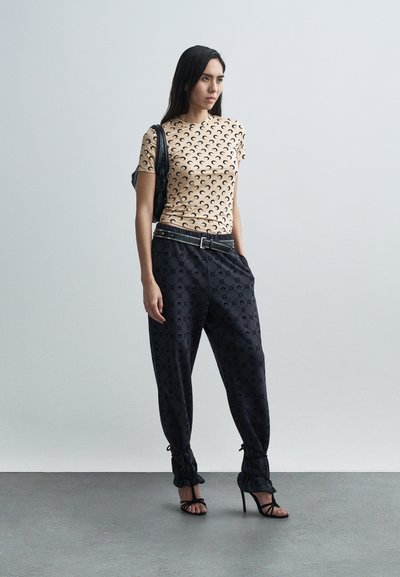 Top beige de manga corta con patrón de medias lunas negras, combinado con pantalones negros con diseño texturizado y lazos en los tobillos, complementados con tacones altos.