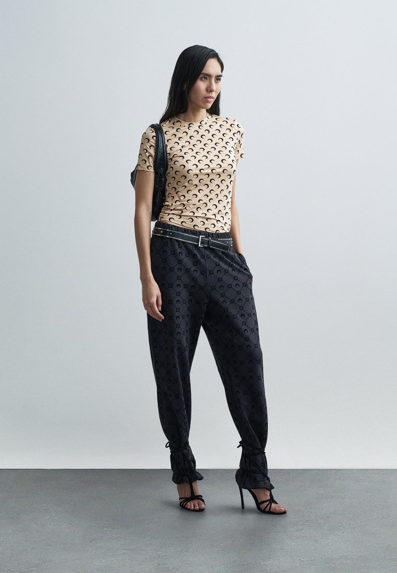 Bézs rövid ujjú felső fekete félhold mintával, fekete texturált mintájú nadrággal és bokán megkötős részletekkel, magas sarkú cipővel kiegészítve.