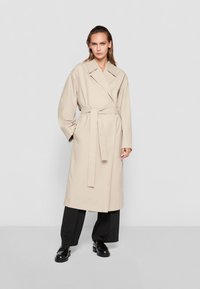 Beige trench coat with a belt, wide lapels, and long sleeves, made of smooth material. Worn over black pants and shoes.
