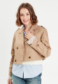 Manteau beige coupé, avec des boutons à double rangée, des épaulettes et une ceinture crème. Associé à une chemise rayée bleue.